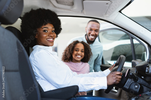 Test drive. Black woman sitting in driver seat of new car, choosing vehicle with husband and daughter at auto dealership