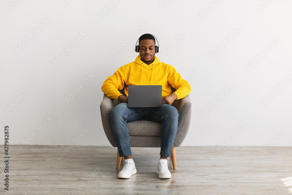 Serious African Guy Using Laptop Wearing Headphones Over Gray Wall Serious African Guy Using Laptop Wearing Headphones Over Gray Wall