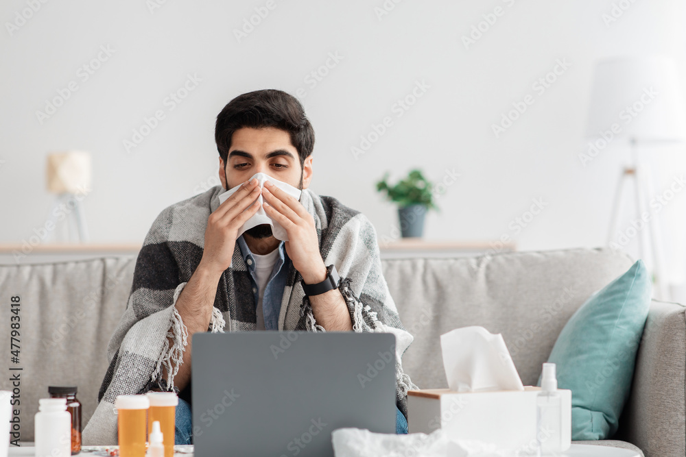 Sick arab man sneezing nose in paper tissue and looking at laptop screen, sitting at home and working, empty space Sick arab man sneezing nose in paper tissue and looking at laptop screen, sitting at home and working, empty space