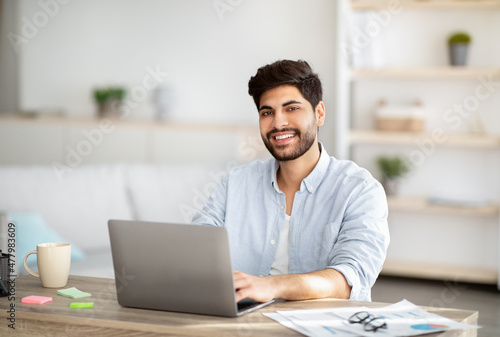 Millennial freelancer. Portrait of happy arab man sitting at desk with laptop, working at home office, copy space