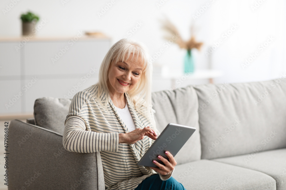 Smiling mature lady using digital tablet sitting on comfy couch Smiling mature lady using digital tablet sitting on comfy couch