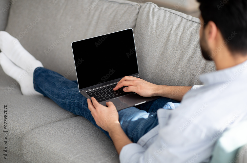 Naklejka premium Young arab guy using modern laptop with empty black screen at home, eclining on couch, mockup, over shoulder shot
