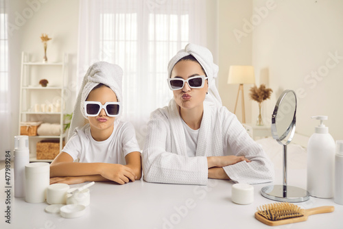 Portrait of beautiful mother and daughter wearing cool trendy shades and white bath towels duck facing sitting at table with mirror and beauty products on spa day or during morning skincare routine