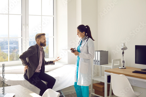 Unhappy young man sitting on examination couch at clinic and telling doctor about his backache symptoms . Male patient talking to physician and asking how to treat bad pain in his injured back photo