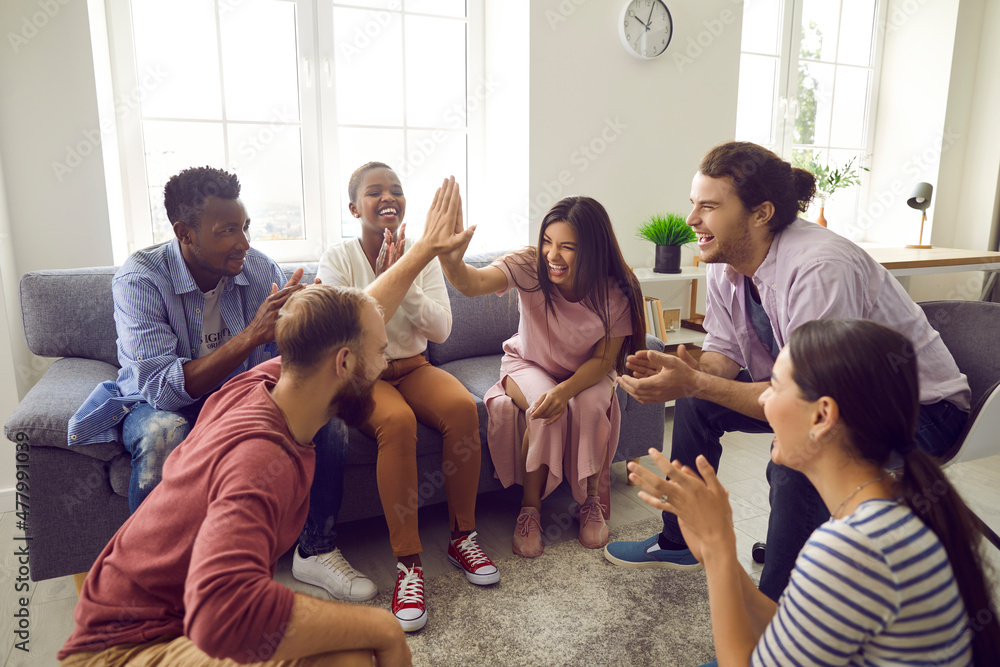 Fototapeta premium Smiling diverse multiracial people have fun give high five relax play together at home. Overjoyed young multiethnic friends celebrate win or victory, engaged in teambuilding. Teamwork concept.