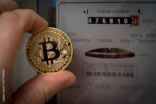 a hand holding a bitcoin coin in front of a three-phase meter photo