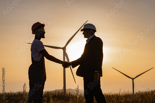 Silhouette of african american engineer and indian inspector using digital tablet during meeting outdoors and shaking hands. Two partners standing on field with wind turbines during amazing sunset. photo