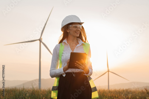 Beautiful caucasian woman in white helmet working with digital tablet at renewable energy farm. Female inspector controlling functioning of wind turbines outdoors. photo