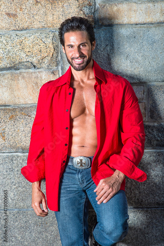 Dressing in a red long sleeve shirt, unbuttoned, and blue jeans, a handsome, sexy, middle age guy with mustache and beard is leaning against the wall, smilingly looking at you.