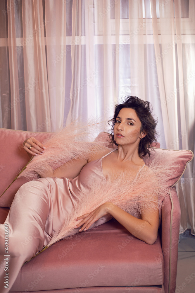 sexy brunette woman in pink dress and feathers lies on the sofa by the window in evening in the room sexy brunette woman in pink dress and feathers lies on the sofa by the window in evening in the room