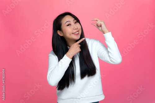 Konstfotografi Happy asian young girl showing with fingers small price little size gesture on pink background
