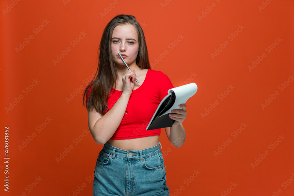 Pensive modern young girl holding notepad and pencil thinking over plans, creative ideas on orange studio background Pensive modern young girl holding notepad and pencil thinking over plans, creative ideas on orange studio background