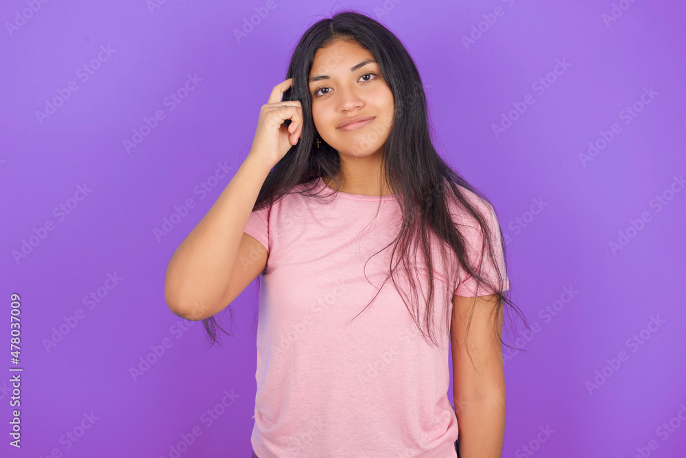 Hispanic brunette girl wearing pink t-shirt over purple background tries to memorize something, keeps fore finger on temple, reminds information for exam, Hispanic brunette girl wearing pink t-shirt over purple background tries to memorize something, keeps fore finger on temple, reminds information for exam,