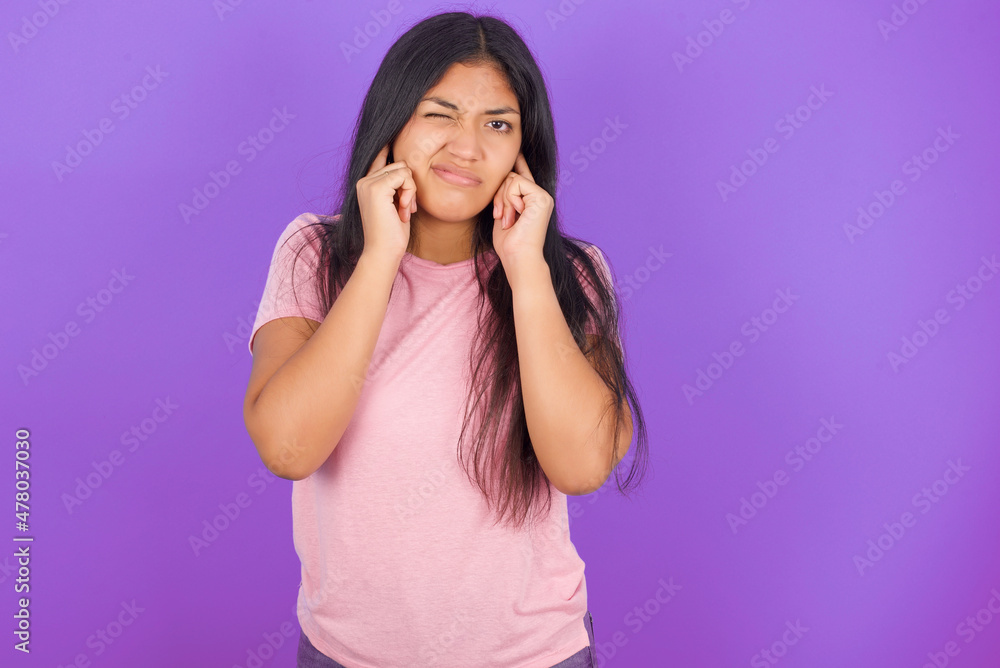 Hispanic brunette girl wearing pink t-shirt over purple background covering ears with fingers with annoyed expression for the noise of loud music. Deaf concept. Hispanic brunette girl wearing pink t-shirt over purple background covering ears with fingers with annoyed expression for the noise of loud music. Deaf concept.