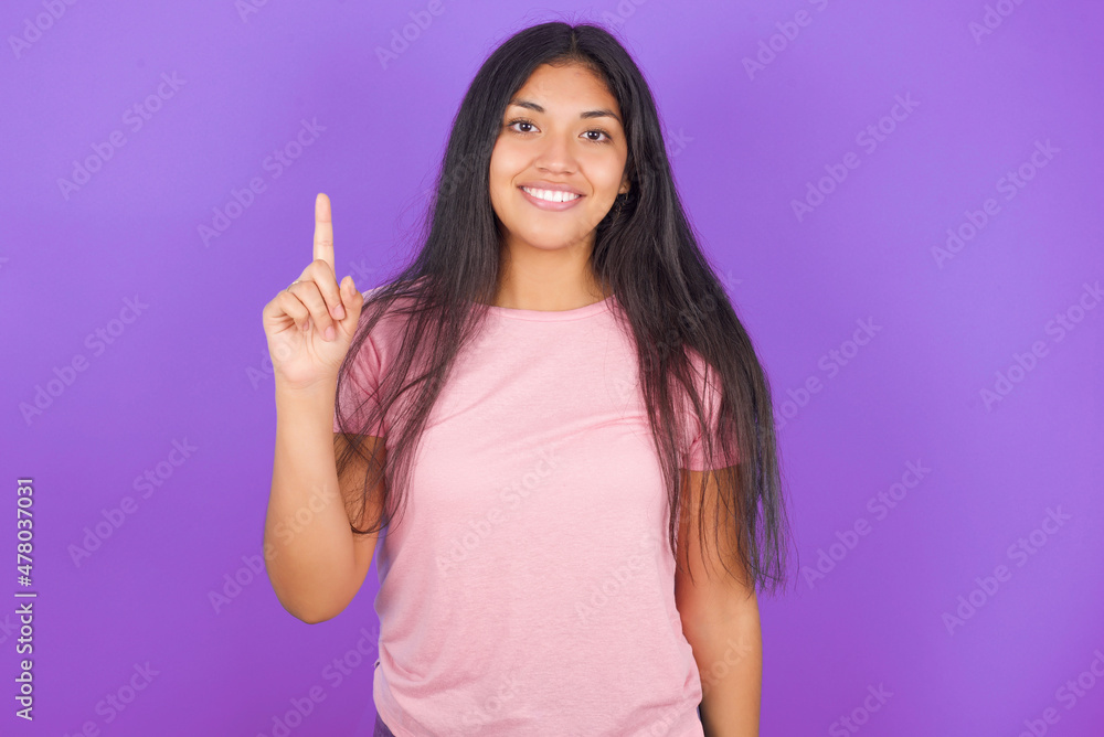 Hispanic brunette girl wearing pink t-shirt over purple background showing and pointing up with finger number one while smiling confident and happy.