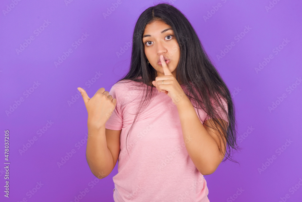 Hispanic brunette girl wearing pink t-shirt over purple background asking to be quiet with finger on lips pointing with hand to the side. Silence and secret concept. Hispanic brunette girl wearing pink t-shirt over purple background asking to be quiet with finger on lips pointing with hand to the side. Silence and secret concept.