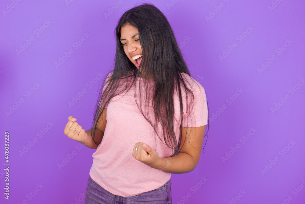 Hispanic brunette girl wearing pink t-shirt over purple background very happy and excited doing winner gesture with arms raised, smiling and screaming for success. Celebration concept. Hispanic brunette girl wearing pink t-shirt over purple background very happy and excited doing winner gesture with arms raised, smiling and screaming for success. Celebration concept.