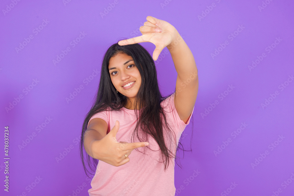 Hispanic brunette girl wearing pink t-shirt over purple background making finger frame with hands. Creativity and photography concept. Hispanic brunette girl wearing pink t-shirt over purple background making finger frame with hands. Creativity and photography concept.