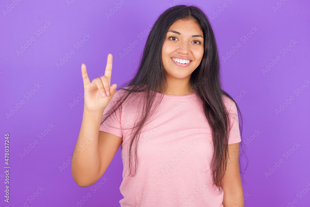 Hispanic brunette girl wearing pink t-shirt over purple background doing a rock gesture and smiling to the camera. Ready to go to her favorite band concert. Hispanic brunette girl wearing pink t-shirt over purple background doing a rock gesture and smiling to the camera. Ready to go to her favorite band concert.
