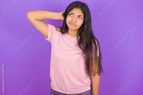 Hispanic brunette girl wearing pink t-shirt over purple background confuse and wonder about question. Uncertain with doubt, thinking with hand on head. Pensive concept.