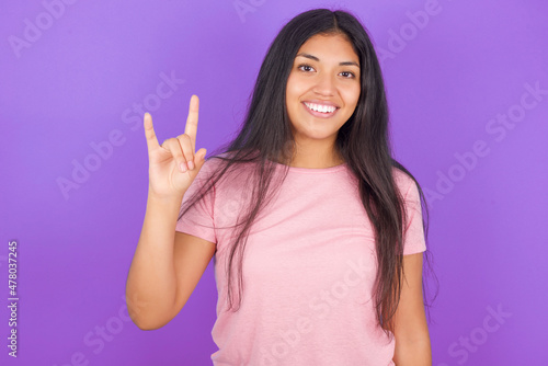 Hispanic brunette girl wearing pink t-shirt over purple background doing a rock gesture and smiling to the camera. Ready to go to her favorite band concert.