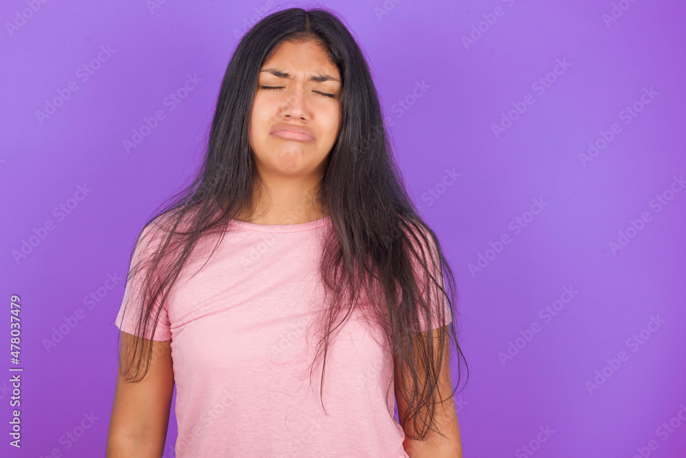 Fototapeta premium Dismal gloomy rejected Hispanic brunette girl wearing pink t-shirt over purple background has problems and difficulties, curves lower lip and closes eyes in despair, being in depression