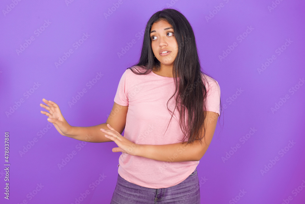 Displeased Hispanic brunette girl wearing pink t-shirt over purple background keeps hands towards empty space and asks not come closer sees something unpleasant Displeased Hispanic brunette girl wearing pink t-shirt over purple background keeps hands towards empty space and asks not come closer sees something unpleasant
