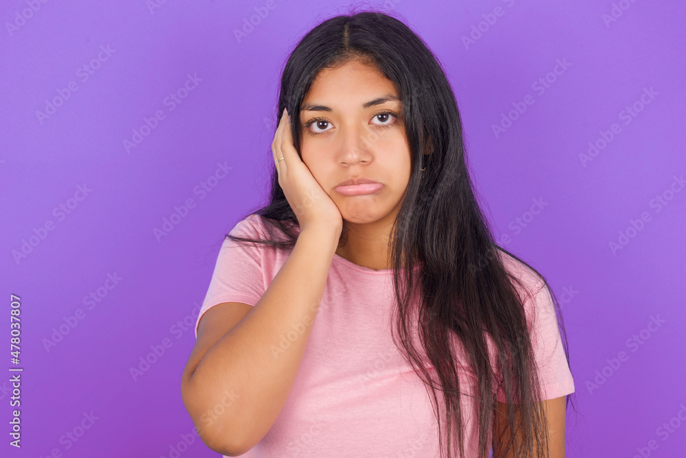 Sad lonely Hispanic brunette girl wearing pink t-shirt over purple background touches cheek with hand bites lower lip and gazes with displeasure. Bad emotions Sad lonely Hispanic brunette girl wearing pink t-shirt over purple background touches cheek with hand bites lower lip and gazes with displeasure. Bad emotions