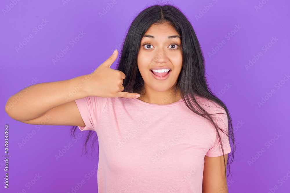 Fototapeta premium Hispanic brunette girl wearing pink t-shirt over purple background makes phone gesture, says call me back again, has glad expression.