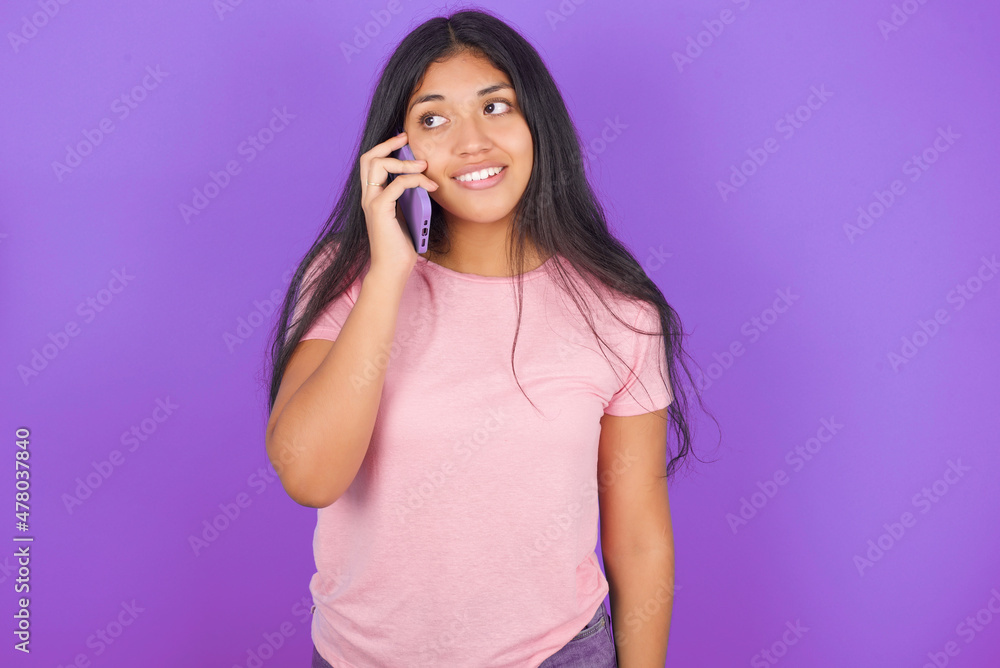 Pleasant looking happy Hispanic brunette girl wearing pink t-shirt over purple backgroundhas nice telephone conversation and looks aside, has nice mood and smiles positively while talks via cell phone Pleasant looking happy Hispanic brunette girl wearing pink t-shirt over purple backgroundhas nice telephone conversation and looks aside, has nice mood and smiles positively while talks via cell phone