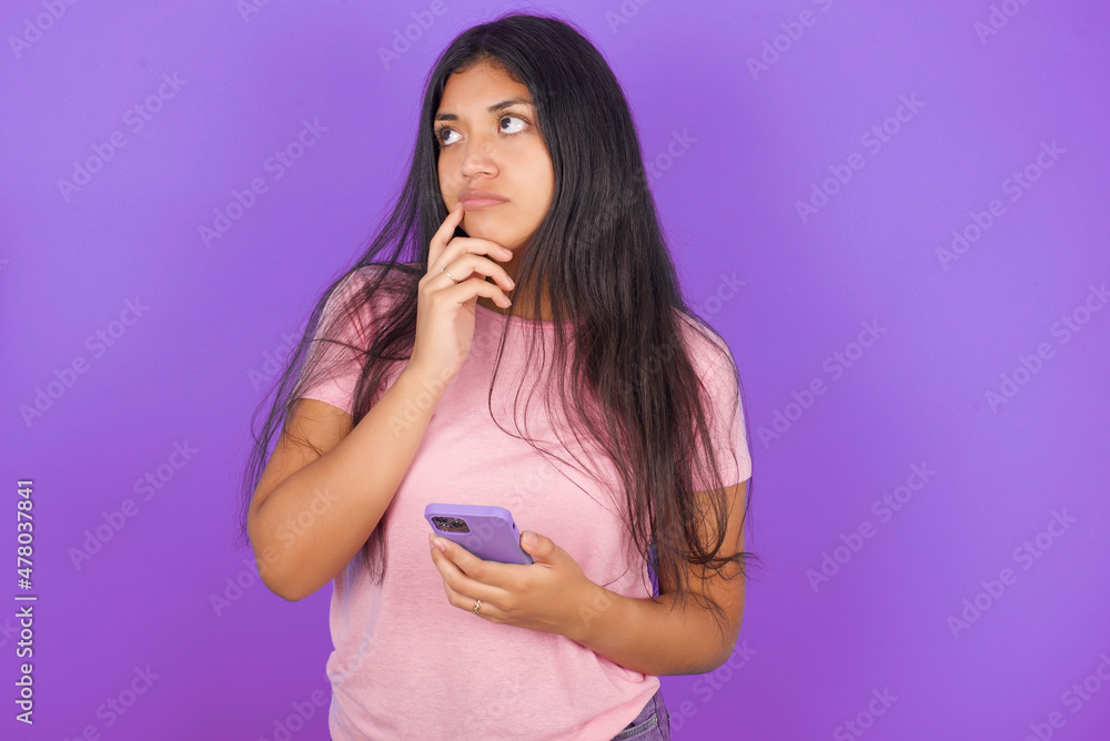 Hispanic brunette girl wearing pink t-shirt over purple background thinks deeply about something, uses modern mobile phone, tries to made up good message, keeps index finger near lips. Hispanic brunette girl wearing pink t-shirt over purple background thinks deeply about something, uses modern mobile phone, tries to made up good message, keeps index finger near lips.