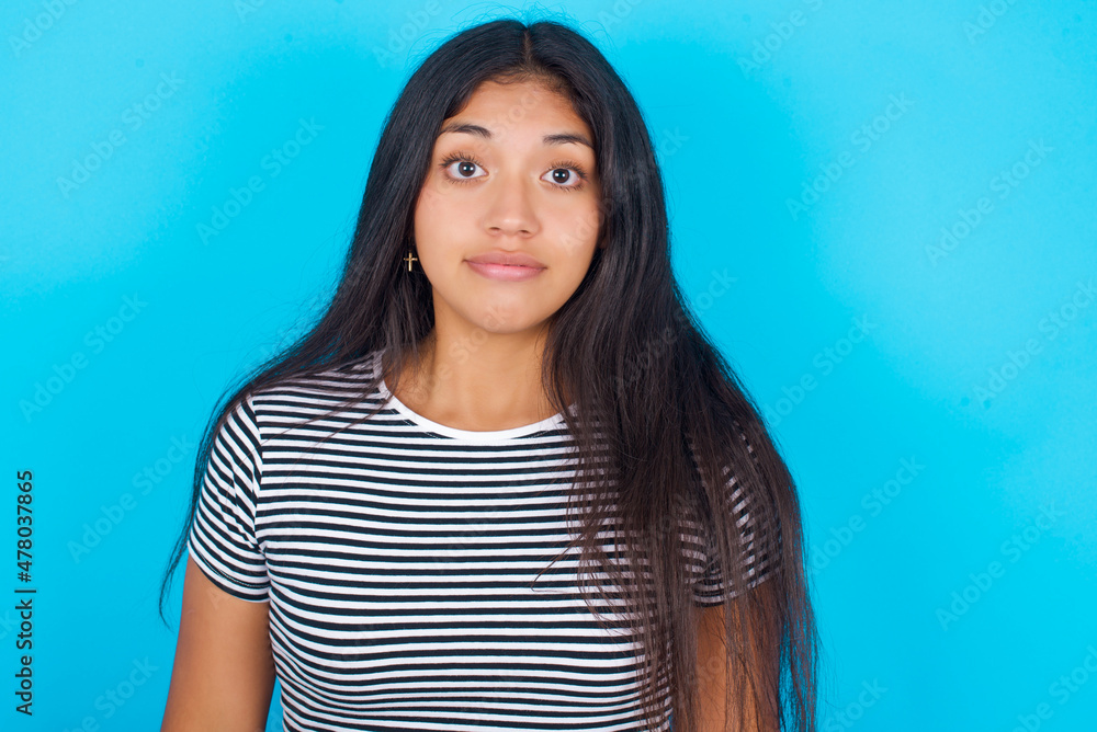 Fototapeta premium Stunned hispanic girl wearing striped t-shirt standing over blue background stares reacts on shocking news. Astonished girl holds breath