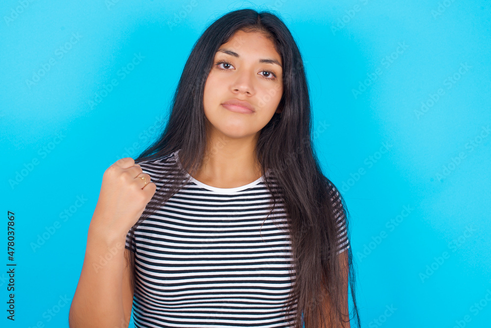 Fototapeta premium hispanic girl wearing striped t-shirt standing over blue background shows fist has annoyed face expression going to revenge or threaten someone makes serious look. I will show you who is boss