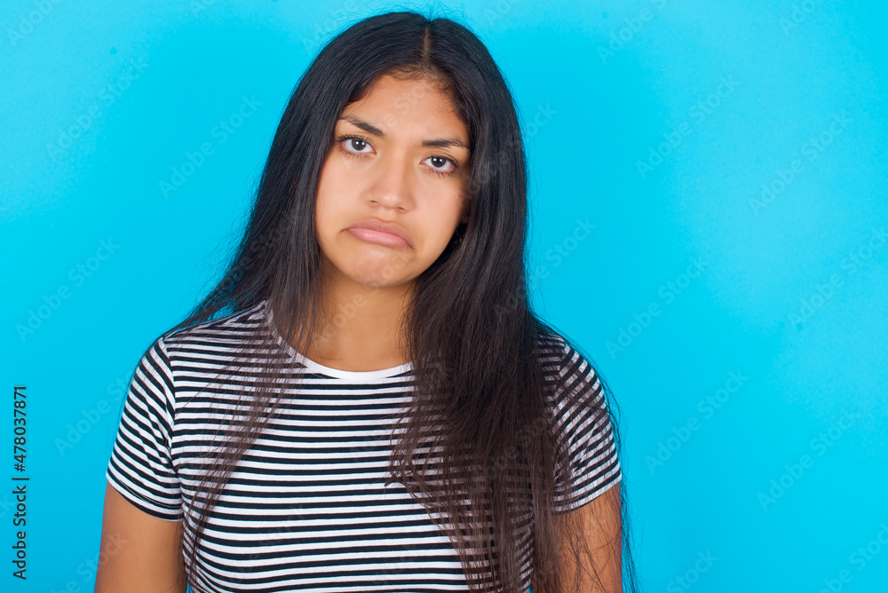 hispanic girl wearing striped t-shirt standing over blue background depressed and worry for distress, crying angry and afraid. Sad expression. hispanic girl wearing striped t-shirt standing over blue background depressed and worry for distress, crying angry and afraid. Sad expression.