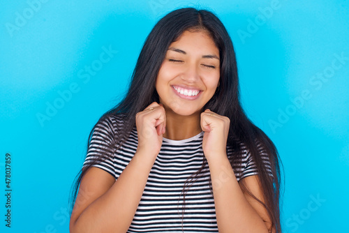 hispanic girl wearing striped t-shirt standing over blue background grins joyfully, imagines something pleasant, copy space. Pleasant emotions concept.