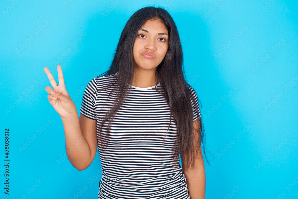 hispanic girl wearing striped t-shirt standing over blue background makes peace gesture keeps lips folded shows v sign. Body language concept hispanic girl wearing striped t-shirt standing over blue background makes peace gesture keeps lips folded shows v sign. Body language concept