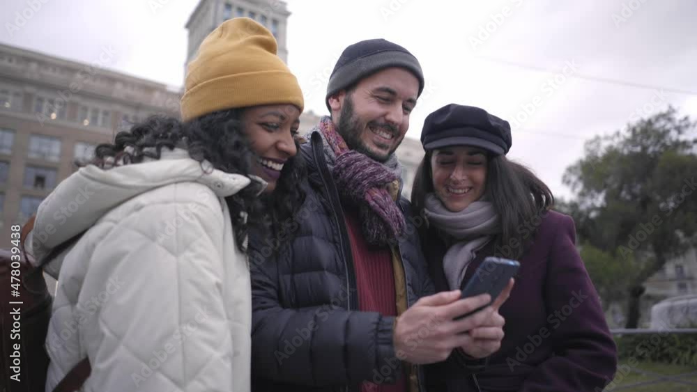 Cheerful people laughing and watching something funny on smartphone. Group of friends in winter clothes using phone outdoors. Holidays makers in a European city. Cheerful people laughing and watching something funny on smartphone. Group of friends in winter clothes using phone outdoors. Holidays makers in a European city.