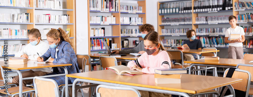 Smart teenagers in face mask spending time in library together and doing their homework.