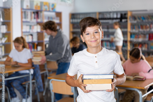 Portrait of a ten-year-old schoolboy standing in the school library, holding textbooks in his hands