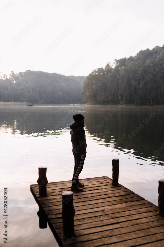 A young man stands alone on a wooden bridge by the lake. Early cloudy foggy morning. To be alone with yourself