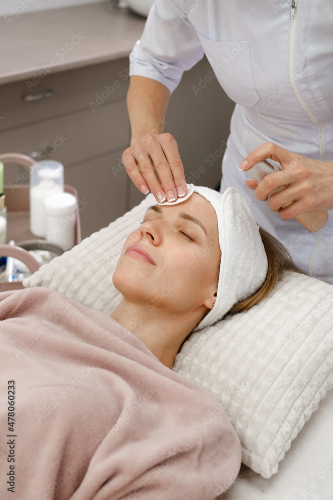 Cosmetologist cleaning skin with cotton pad, care and treatment procedure in modern aesthetic clinic. Woman applies tonic on face in beauty salon