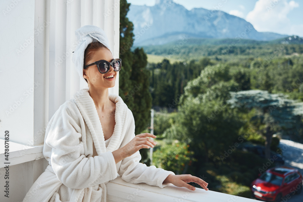 Portrait of gorgeous woman wearing sunglasses posing in a bathrobe on a balcony rest Perfect sunny morning