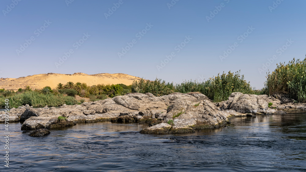 Picturesque boulders lie on the riverbank, green vegetation is visible. Sand dunes against the azure sky. Ripples on the blue water. Egypt. Nile