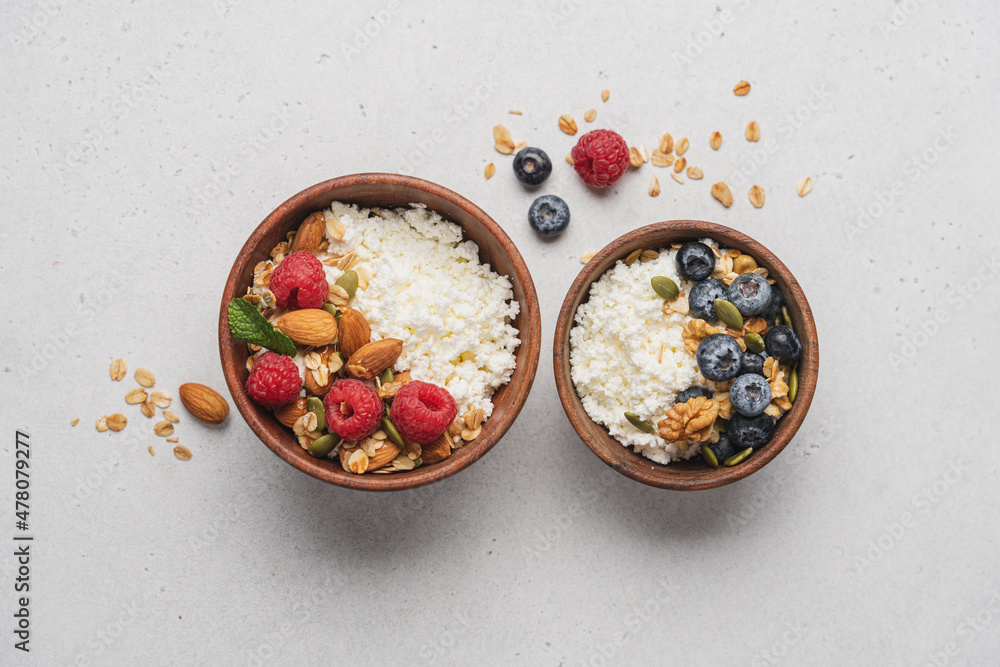 Fototapeta premium Two different Cottage cheese bowl with blueberries and raspberries, granola, almonds, walnuts and honey. Servings of healthy breakfast on light gray background
