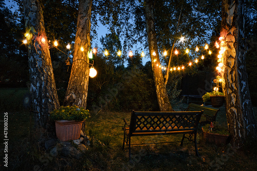 Fototapeta Naklejka Na Ścianę i Meble -  string of lights in the garden with garden bench and sheltered corner hidden by hedges in the evening.