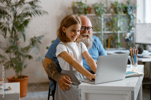Joyful little girl with father do hometask on laptop together sitting at table photo