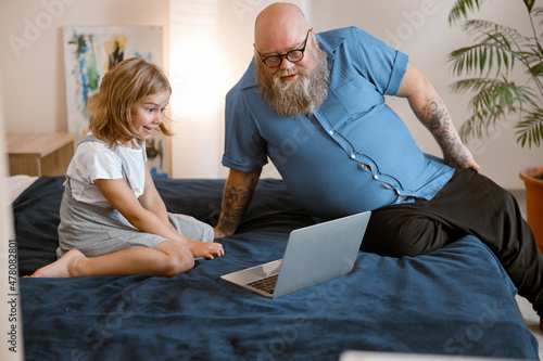 Bearded father and little girl watch moovie lesson via modern laptop sitting on bed photo