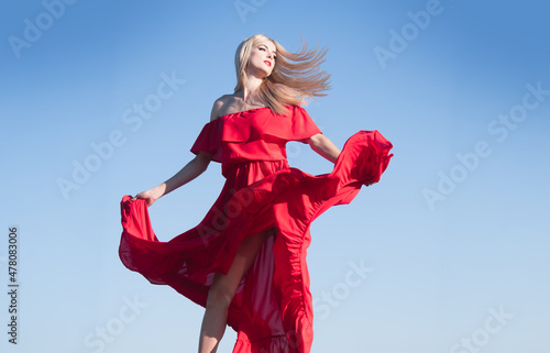 Blonde woman in red dress movement on sky. Fashionable young model in style dress. Fashion woman in summer.