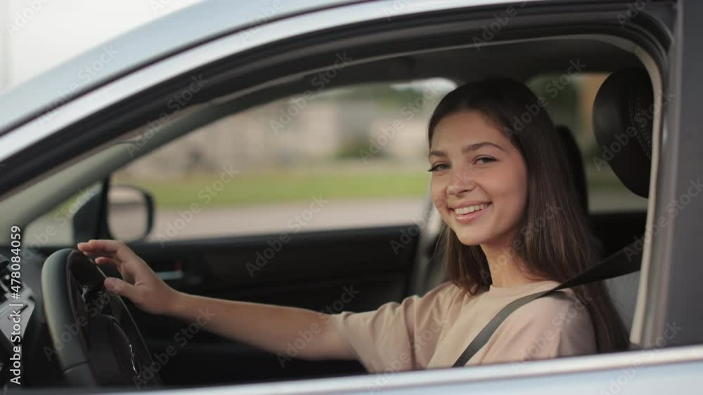 A young woman is sitting in a car and wearing a seat belt. She is smiling and looking at the camera. She has her hand on the steering wheel. 4K A young woman is sitting in a car and wearing a seat belt. She is smiling and looking at the camera. She has her hand on the steering wheel. 4K