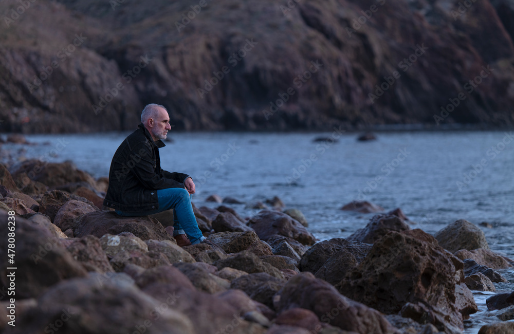 Adult man in black jacket sitting on rocky beach looking at sea. Almeria, Spain